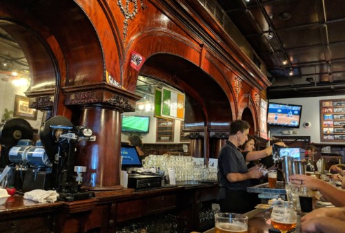 A lively bar scene with wooden decor, patrons enjoying drinks, and screens showing sports events.