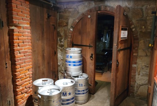 A rustic cellar entrance with wooden doors and stacked beer kegs in front of a stone wall.