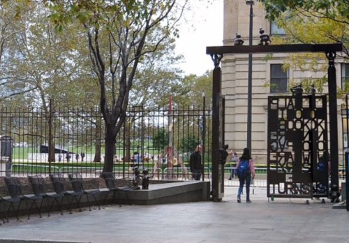 A park entrance with a decorative gate, benches, and trees, with people walking in the background.