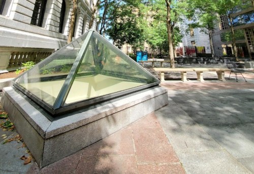 A glass pyramid structure in a park, surrounded by trees and benches, with a building in the background.