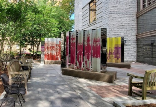 Outdoor area featuring colorful glass panels and benches, surrounded by greenery and a stone building.