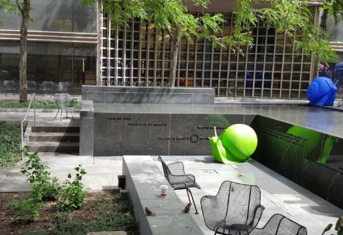 A modern outdoor space with green and blue sculptures, seating, and a reflective water feature surrounded by trees.