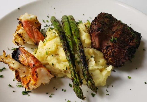 A plate featuring grilled shrimp, asparagus, and a portion of creamy mashed potatoes with a seasoned meat cut.