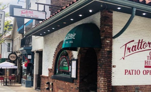 Exterior of Trattoria on the Hill restaurant, featuring a green awning and brick facade, with outdoor seating nearby.