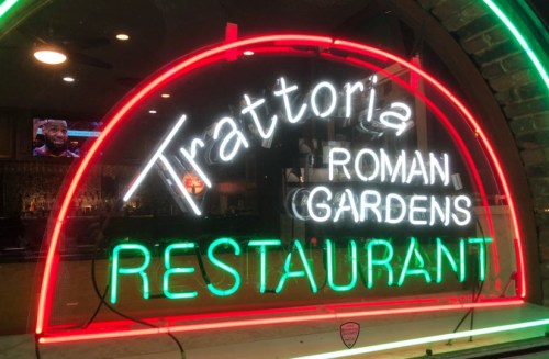 Neon sign for "Trattoria Roman Gardens Restaurant" in vibrant green and white lights.