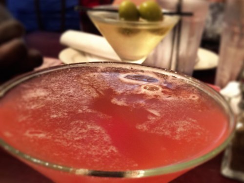 Two cocktails on a table: a pink drink in the foreground and a martini with olives in the background.