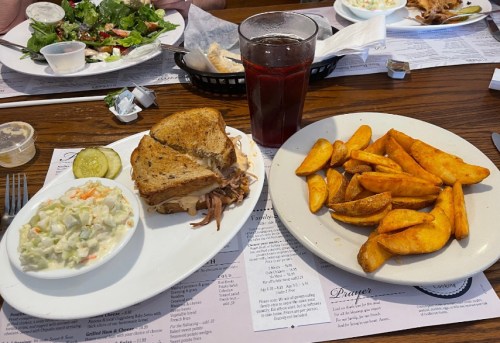 A plate with a pulled pork sandwich, coleslaw, and a side of potato wedges, alongside a glass of iced tea.