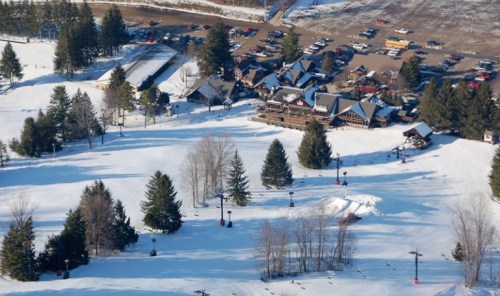 A snowy landscape with ski slopes, trees, and a lodge surrounded by parked cars in a winter setting.