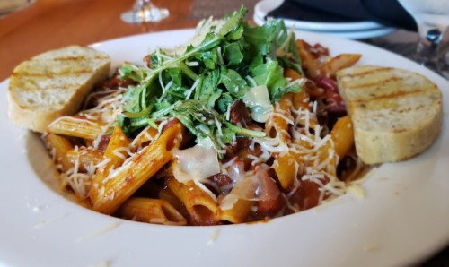 A plate of pasta with meat sauce, topped with cheese and arugula, served with two slices of grilled garlic bread.