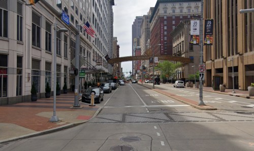 A wide city street lined with buildings, parked cars, and a decorative arch overhead, under a cloudy sky.