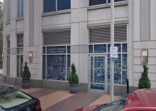 Exterior view of a building featuring a "Happy Hour" sign and the entrance to Ghostlight, with planters and parked cars.