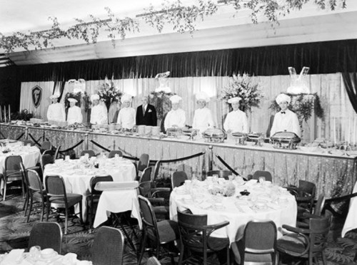 A vintage banquet scene with chefs and a lavish buffet, surrounded by elegantly set tables in a decorated hall.