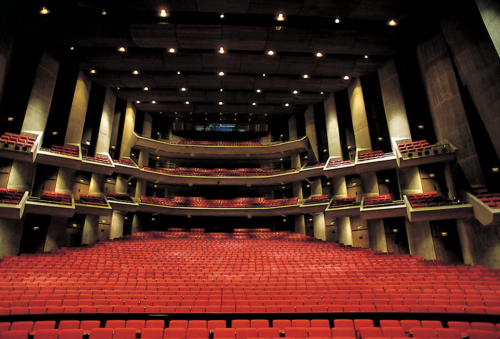 Interior of a theater with multiple levels of red seating and a spacious stage area, illuminated by overhead lights.