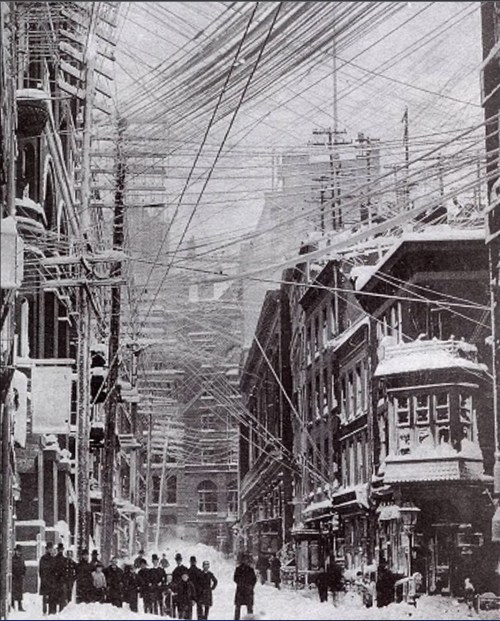 Historic street scene with snow, featuring tangled telegraph wires overhead and people walking in the foreground.
