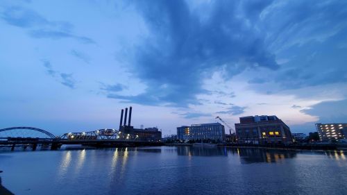 A serene evening view of a river with buildings and smokestacks under a cloudy blue sky.