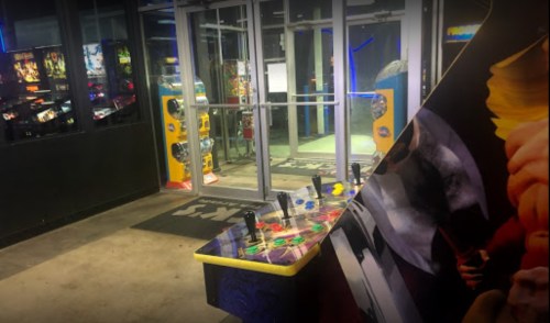 A dimly lit arcade entrance with a colorful game cabinet in the foreground and glass doors leading outside.