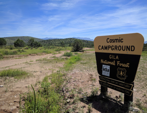 Sign for Cosmic Campground in Gila National Forest, surrounded by greenery and mountains under a clear blue sky.