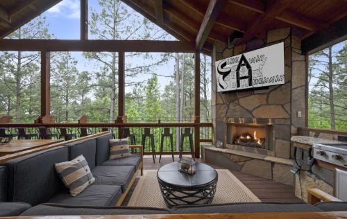 Cozy cabin interior with a stone fireplace, modern seating, and large windows overlooking a forested view.