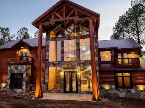 A modern log cabin with large glass windows and stone accents, surrounded by trees at dusk.