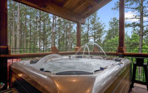 A hot tub on a wooden deck surrounded by tall trees and a clear blue sky. Water jets create a relaxing atmosphere.