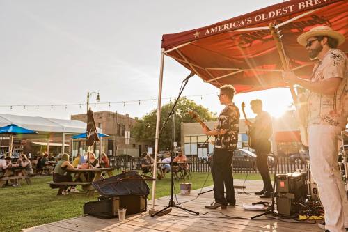 A live band performs on stage at an outdoor venue during sunset, with an audience seated at picnic tables.