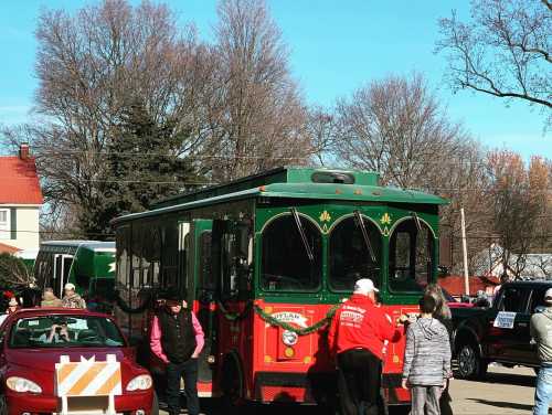 A festive green and red trolley parked on a street, surrounded by people and trees on a sunny day.