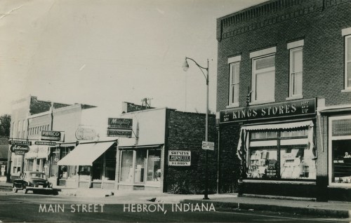 Black and white photo of Main Street in Hebron, Indiana, featuring storefronts and a vintage car parked nearby.
