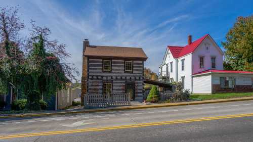 A log cabin and a white house with a red roof sit side by side along a quiet street under a blue sky.