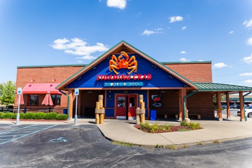 Exterior of a seafood restaurant with a colorful crab sign, featuring a red roof and outdoor seating area.