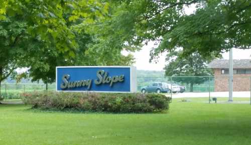 Sign for "Sunny Slope" surrounded by greenery, with a building and cars visible in the background.