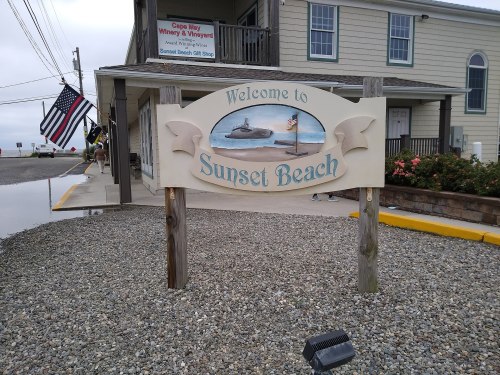 Sign reading "Welcome to Sunset Beach" in front of a building, with an American flag and gravel foreground.