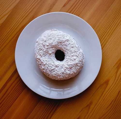 A powdered sugar-covered donut on a white plate, resting on a wooden table.