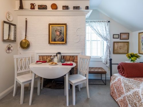 Cozy room with a white table, two chairs, a vintage painting, and a small fireplace, decorated with flowers and plates.