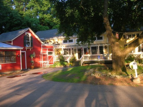 A charming scene of a red and yellow house with a porch, surrounded by greenery and a large tree in the foreground.