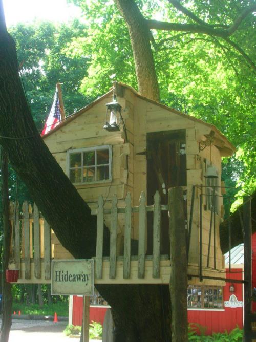 A wooden treehouse with a small porch, surrounded by trees, featuring a lantern and an American flag. Sign reads "Hideaway."