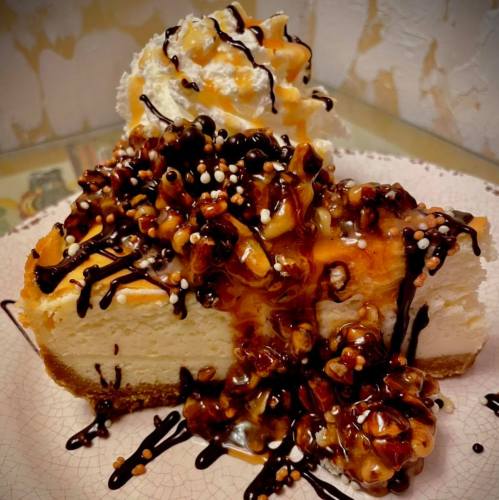 A slice of cheesecake topped with caramel, chocolate drizzle, nuts, and whipped cream on a decorative plate.