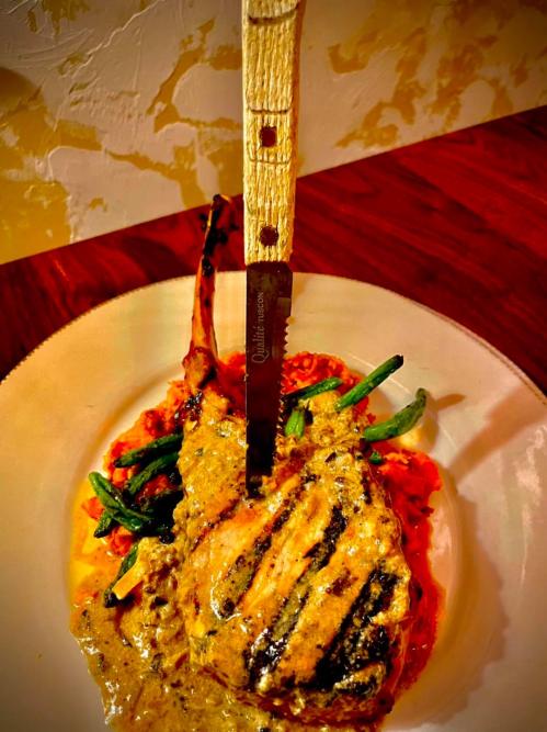 A grilled pork chop with a knife resting in it, served on a plate with green beans and a creamy sauce.