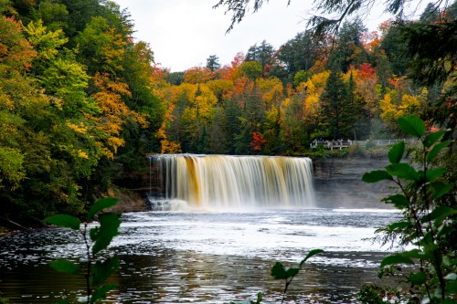 A serene waterfall cascades into a river, surrounded by vibrant autumn foliage and lush greenery.