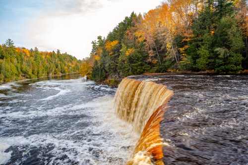 A waterfall cascades into a river, surrounded by autumn foliage and lush green trees under a clear sky.