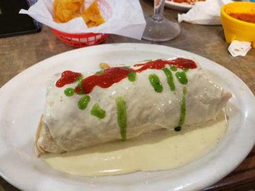A large burrito topped with red and green sauces, served on a plate with a creamy sauce. Chips in the background.