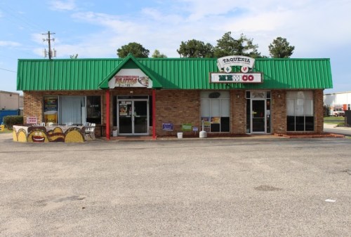 A brick building with a green roof, featuring a taqueria and a restaurant, surrounded by a parking lot.