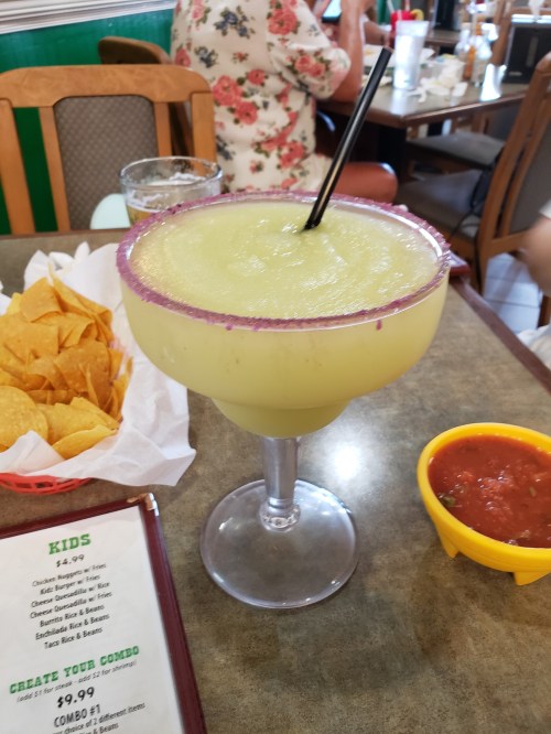 A large margarita with a purple rim and a straw, next to a plate of tortilla chips and salsa on a restaurant table.
