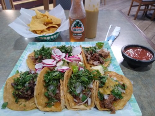 A plate of tacos topped with radishes and cilantro, served with chips, salsa, and a bottle of Jarritos soda.