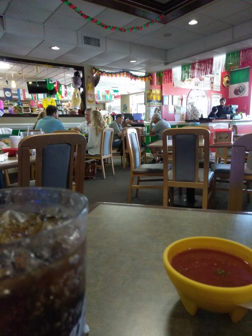 A busy restaurant interior with colorful decorations, tables filled with diners, and a glass of soda in the foreground.