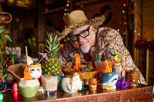 A man in a straw hat and floral shirt smiles behind a colorful display of tropical cocktails and decorations.