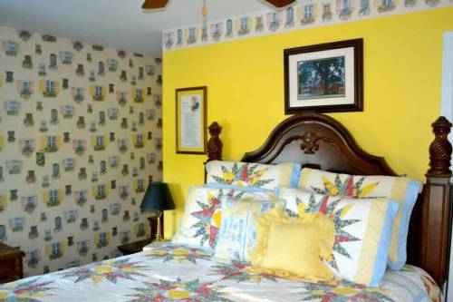 Cozy bedroom with a yellow wall, patterned wallpaper, a wooden bed, and a colorful quilt. Decor includes framed art.