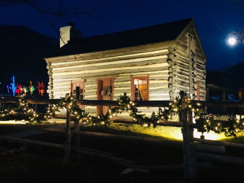 A cozy log cabin illuminated at night, surrounded by festive lights and a full moon in the sky.