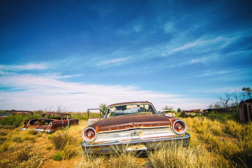 Abandoned vintage cars rusting in a field under a bright blue sky with scattered clouds.