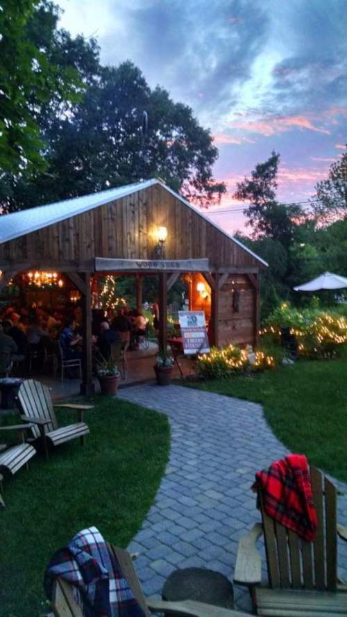 A cozy wooden building with outdoor seating, surrounded by greenery and twinkling lights, at sunset.