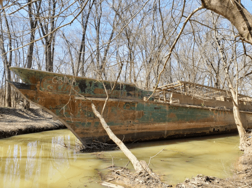 An abandoned, rusted boat partially submerged in a muddy river, surrounded by bare trees.